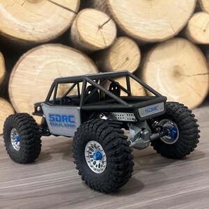 K&ouml;nnte beinhalten: Ein grauer und schwarzer ferngesteuerter Crawler mit gro&szlig;en, schwarzen Reifen und blauen Akzenten. Das Fahrzeug hat einen &Uuml;berrollk&auml;fig und der Text "SDRC Crawl Shop" ist sichtbar.