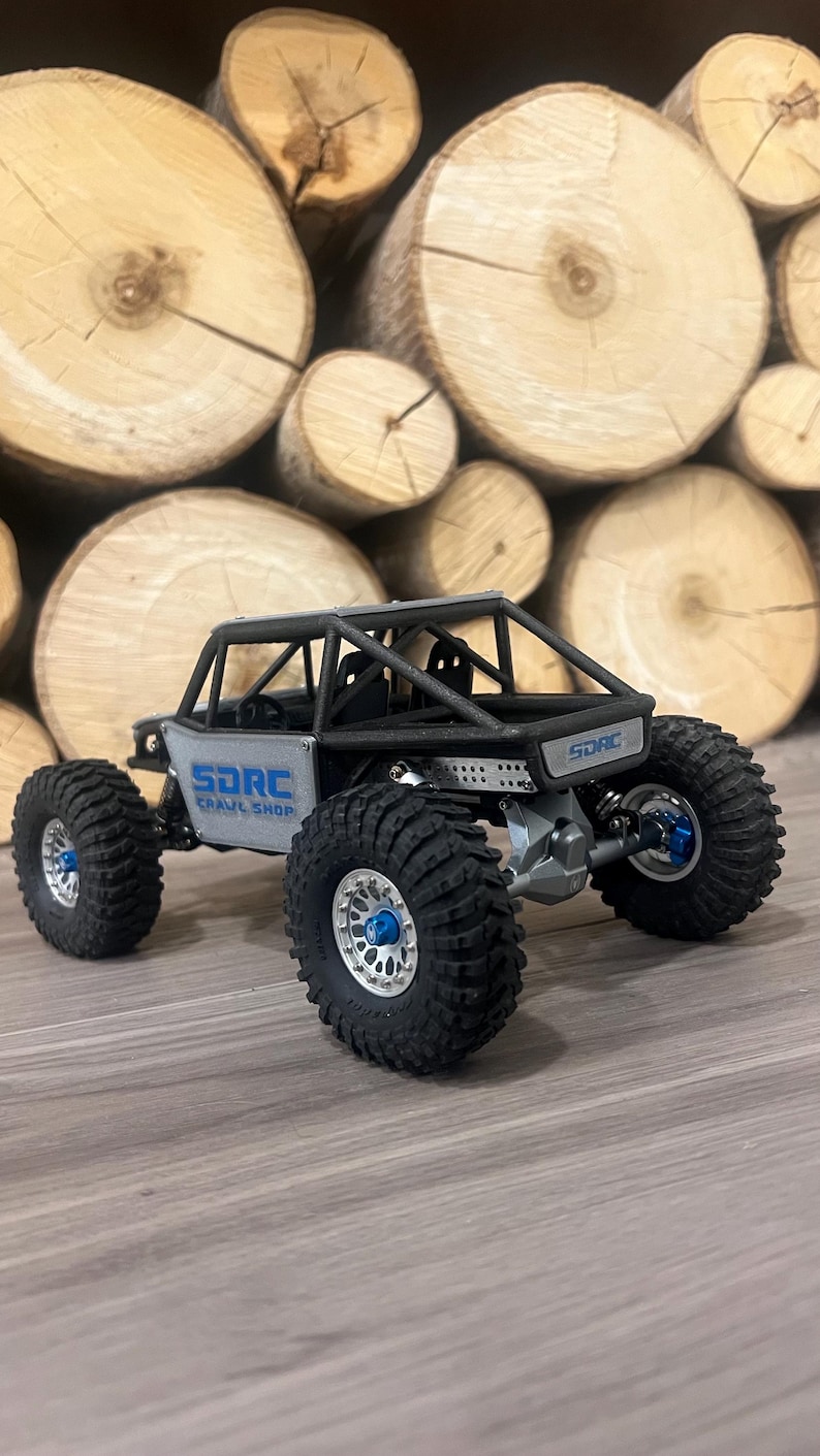 K&ouml;nnte beinhalten: Ein grauer und schwarzer ferngesteuerter Crawler mit gro&szlig;en, schwarzen Reifen und blauen Akzenten. Das Fahrzeug hat einen &Uuml;berrollk&auml;fig und der Text "SDRC Crawl Shop" ist sichtbar.