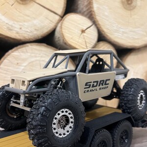 K&ouml;nnte beinhalten: Ein beigefarbener und grauer ferngesteuerter Crawler-Truck mit gro&szlig;en, schwarzen Offroad-Reifen wird auf einem schwarzen Anh&auml;nger gezeigt. Der Truck hat einen &Uuml;berrollk&auml;fig und den Text "SDRC CRAWL SHOP" an der Seite. Der Hintergrund zeigt einen Stapel geschnittener Baumst&auml;mme.