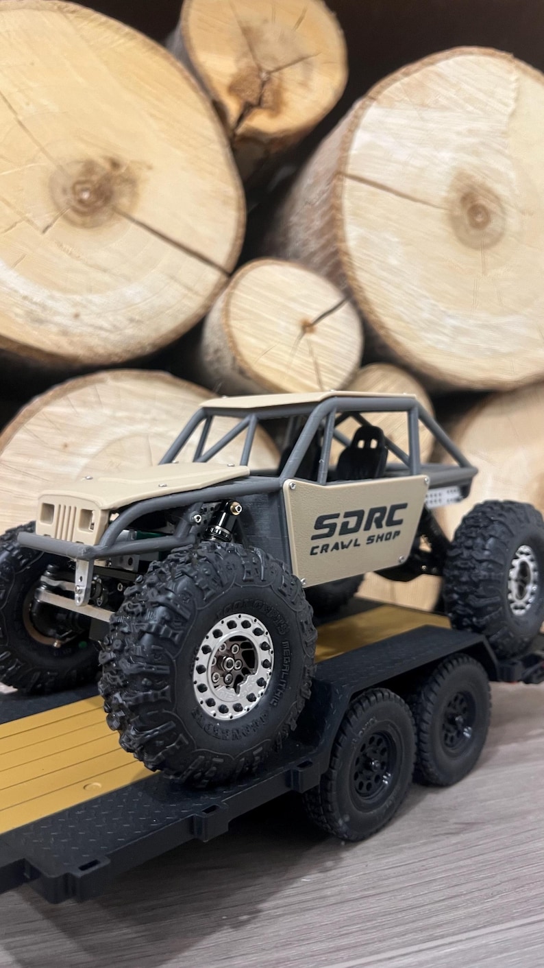 K&ouml;nnte beinhalten: Ein beigefarbener und grauer ferngesteuerter Crawler-Truck mit gro&szlig;en, schwarzen Offroad-Reifen wird auf einem schwarzen Anh&auml;nger gezeigt. Der Truck hat einen &Uuml;berrollk&auml;fig und den Text "SDRC CRAWL SHOP" an der Seite. Der Hintergrund zeigt einen Stapel geschnittener Baumst&auml;mme.