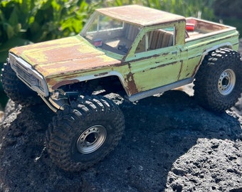 May include: A rusty green toy truck with large tires, sitting on a dark gray rock. The truck has a white painted trim and a black interior.