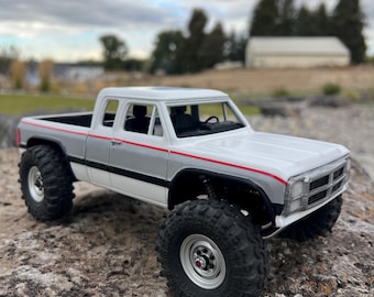 May include: A white and gray model pickup truck with large black tires. The truck has a red stripe and is sitting on a rock. The background shows a cloudy sky and trees.