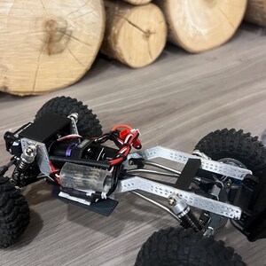 K&ouml;nnte beinhalten: Ein detailliertes, teilweise montiertes ferngesteuertes Crawler-Truck in Schwarz und Silber. Das Fahrzeug verf&uuml;gt &uuml;ber gro&szlig;e, grobstollige Reifen, ein komplexes Federungssystem und freiliegende mechanische Komponenten. Der Hintergrund zeigt gestapelte Holzscheite.