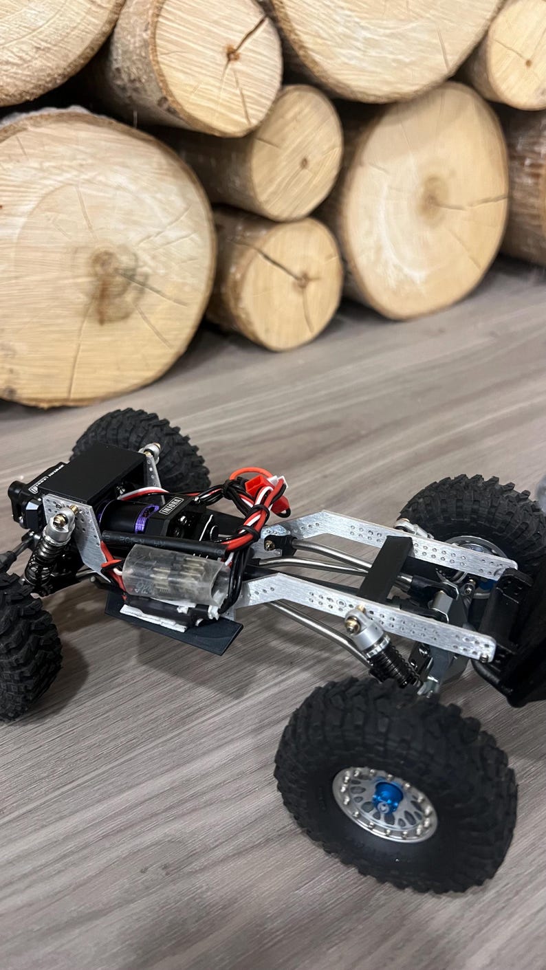 K&ouml;nnte beinhalten: Ein detailliertes, teilweise montiertes ferngesteuertes Crawler-Truck in Schwarz und Silber. Das Fahrzeug verf&uuml;gt &uuml;ber gro&szlig;e, grobstollige Reifen, ein komplexes Federungssystem und freiliegende mechanische Komponenten. Der Hintergrund zeigt gestapelte Holzscheite.