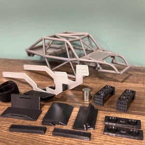K&ouml;nnte beinhalten: Ein zerlegter Modellauto-Bausatz mit verschiedenen Komponenten. Das Set enth&auml;lt einen grauen &Uuml;berrollk&auml;fig, schwarze und graue Kunststoffteile sowie einen kleinen Metallzylinder. Die Teile sind auf einer Holzoberfl&auml;che angeordnet.