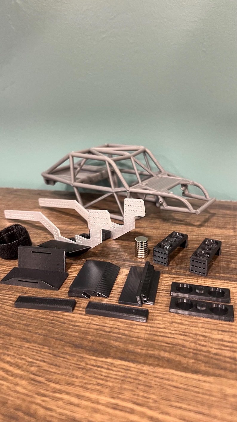 K&ouml;nnte beinhalten: Ein zerlegter Modellauto-Bausatz mit verschiedenen Komponenten. Das Set enth&auml;lt einen grauen &Uuml;berrollk&auml;fig, schwarze und graue Kunststoffteile sowie einen kleinen Metallzylinder. Die Teile sind auf einer Holzoberfl&auml;che angeordnet.