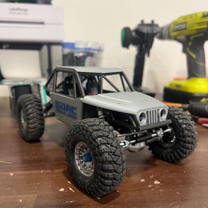 K&ouml;nnte beinhalten: Ein grauer und schwarzer RC-Rock-Crawler mit gro&szlig;en, grobstolligen Reifen. Das Modell hat den Text "SDRC" auf der Seite. Im Hintergrund sind ein gr&uuml;nes RC-Auto und Werkzeuge zu sehen.