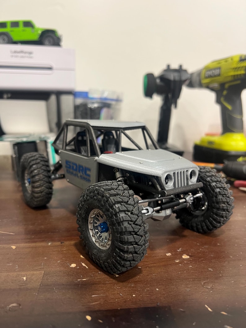 K&ouml;nnte beinhalten: Ein grauer und schwarzer RC-Rock-Crawler mit gro&szlig;en, grobstolligen Reifen. Das Modell hat den Text "SDRC" auf der Seite. Im Hintergrund sind ein gr&uuml;nes RC-Auto und Werkzeuge zu sehen.
