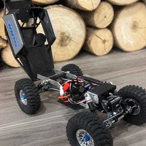 K&ouml;nnte beinhalten: Ein schwarzer und silberner ferngesteuerter Rock Crawler mit gro&szlig;en, grobstolligen Reifen. Das Fahrzeug hat einen &Uuml;berrollk&auml;fig und sichtbare interne Komponenten, einschlie&szlig;lich Verkabelung und Motor. Die R&auml;der haben blaue Akzente.