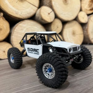 K&ouml;nnte beinhalten: Ein wei&szlig;-schwarzes ferngesteuertes Crawler-Modell mit gro&szlig;en, schwarzen Reifen und silbernen Felgen. Das Fahrzeug hat einen &Uuml;berrollk&auml;fig und den Text "SDRC CRAWL SHOP" an der Seite. Der Hintergrund zeigt einen Stapel Holzscheite.