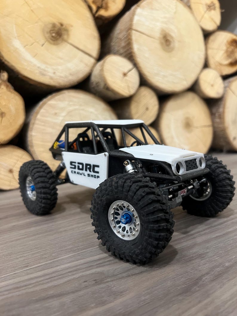 K&ouml;nnte beinhalten: Ein wei&szlig;-schwarzes ferngesteuertes Crawler-Modell mit gro&szlig;en, schwarzen Reifen und silbernen Felgen. Das Fahrzeug hat einen &Uuml;berrollk&auml;fig und den Text "SDRC CRAWL SHOP" an der Seite. Der Hintergrund zeigt einen Stapel Holzscheite.