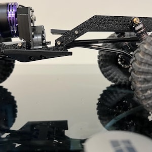 May include: A black and silver 3D printed model of a truck chassis with a purple motor and black wheels. The chassis is on a black surface with a white label that says "Bambu Lab".