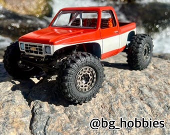 May include: A red and white custom-painted truck with a roll cage and large off-road tires. The truck is sitting on a rocky surface. @bg_hobbies
