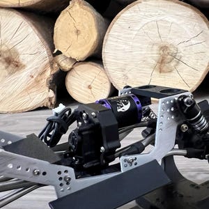 May include: Close-up of a black and silver RC car chassis with a purple motor and black components. The background features a stack of light brown wooden logs.