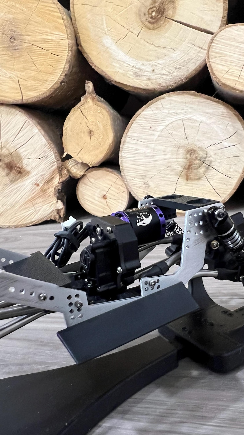 May include: Close-up of a black and silver RC car chassis with a purple motor and black components. The background features a stack of light brown wooden logs.