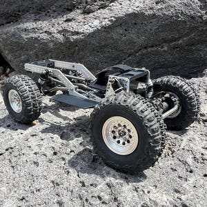 May include: A silver and black remote control rock crawler with large, knobby tires. The vehicle has a detailed chassis and suspension system, designed for off-road terrain. The crawler is positioned on a rocky surface.