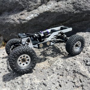 May include: A small, detailed, black and silver remote control truck with large, knobby tires. The truck has a complex suspension system and is positioned on a rough, gray rock surface. The wheels are silver with black tires.