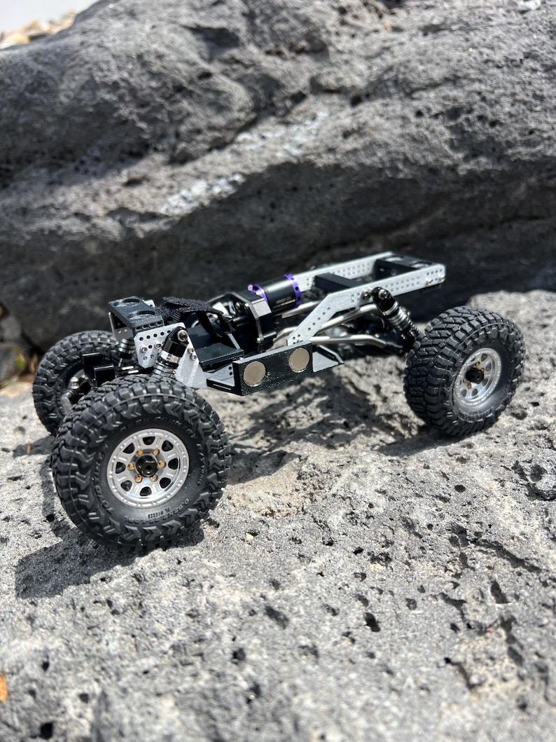 May include: A small, detailed, black and silver remote control truck with large, knobby tires. The truck has a complex suspension system and is positioned on a rough, gray rock surface. The wheels are silver with black tires.