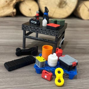 May include: Miniature workshop scene featuring a black metal table with various tools and equipment. Includes a black toolbox, orange container, red gas can, and a yellow figure eight. Other items include a battery, a first aid kit, and a small black vehicle.