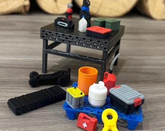 May include: Miniature workshop scene featuring a black metal table with various tools and equipment. Includes a black toolbox, orange container, red gas can, and a yellow figure eight. Other items include a battery, a first aid kit, and a small black vehicle.