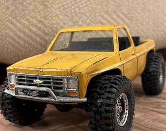 Op de afbeelding: Een gele speelgoed pick-up truck met grote zwarte banden. De truck heeft een zilveren voorbumper en een zwarte laadbak. De truck is een miniatuur replica van een klassiek model.