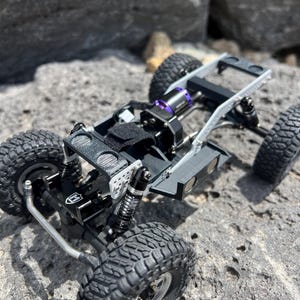 May include: A detailed view of a small, black and silver remote control truck chassis with large, textured tires. The chassis features a black motor with purple accents and is set against a rocky background.