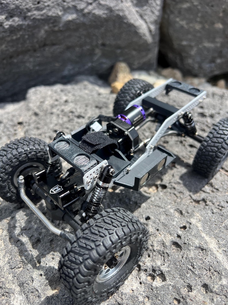 May include: A detailed view of a small, black and silver remote control truck chassis with large, textured tires. The chassis features a black motor with purple accents and is set against a rocky background.