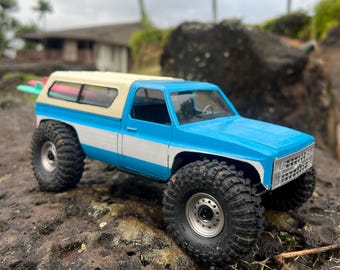 Peut inclure: Un camion jouet bleu et blanc avec un toit blanc et une planche de surf à l'arrière. Le camion est sur une surface rocheuse avec de l'herbe verte et des palmiers en arrière-plan.