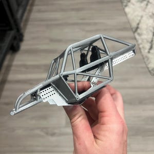K&ouml;nnte beinhalten: Ein graues Modellauto-Chassis mit &Uuml;berrollk&auml;fig, Sitzen und perforierten Metallplatten. Das Miniaturfahrzeug wird in einer Hand gehalten, was sein detailliertes Design zeigt. Das Chassis scheint ein Modell eines Buggys oder Gel&auml;ndewagens zu sein.