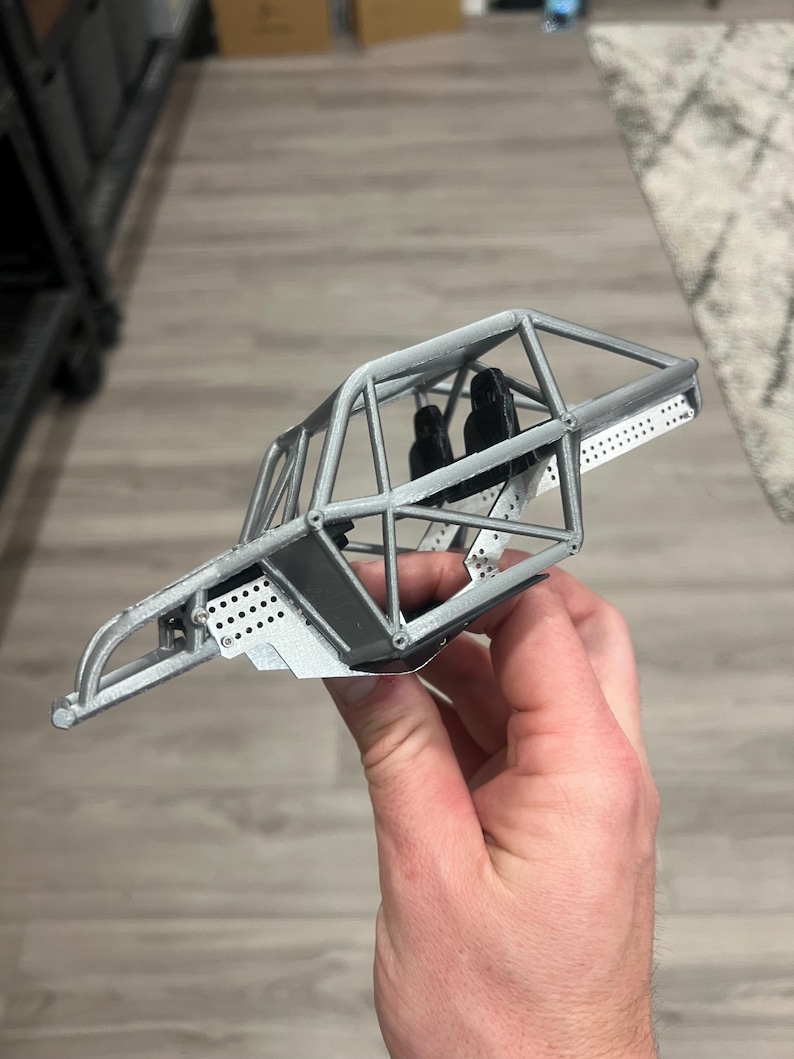 K&ouml;nnte beinhalten: Ein graues Modellauto-Chassis mit &Uuml;berrollk&auml;fig, Sitzen und perforierten Metallplatten. Das Miniaturfahrzeug wird in einer Hand gehalten, was sein detailliertes Design zeigt. Das Chassis scheint ein Modell eines Buggys oder Gel&auml;ndewagens zu sein.