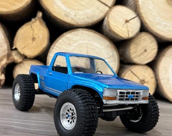 May include: A blue toy truck with black tires and silver wheels. The truck is sitting on a wooden surface in front of a stack of logs.