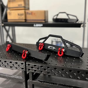 May include: Black metal off-road vehicle bumpers with red tow hooks. The bumpers are displayed on a black metal surface. A shelf in the background holds tires and boxes. The word "LBRP" is visible on the shelf.