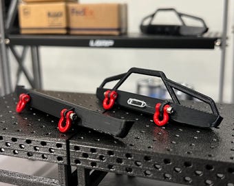 May include: Black metal off-road vehicle bumpers with red tow hooks. The bumpers are displayed on a black metal surface. A shelf in the background holds tires and boxes. The word "LBRP" is visible on the shelf.