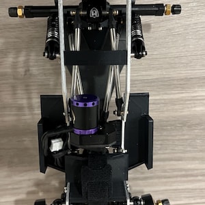 May include: A black and silver RC car chassis with a purple motor and gold-colored accents. The chassis has a complex design with multiple suspension components, including springs and shocks. Two silver magnets are visible on the front and top.