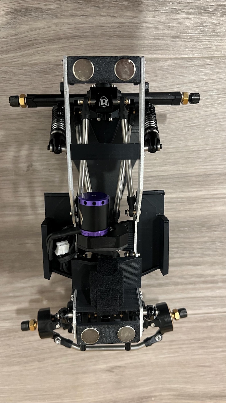 May include: A black and silver RC car chassis with a purple motor and gold-colored accents. The chassis has a complex design with multiple suspension components, including springs and shocks. Two silver magnets are visible on the front and top.
