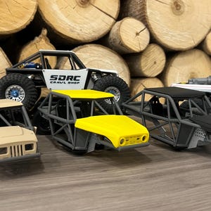 K&ouml;nnte beinhalten: Mehrere Miniatur-Gel&auml;ndewagen werden gezeigt, darunter ein wei&szlig;-schwarzer Rock Crawler mit dem Text "SDRC Crawl Shop", ein gelbbedachtes Buggy und ein wei&szlig;er Pick-up. Die Fahrzeuge stehen vor einem Hintergrund aus gestapelten Holzscheiten.