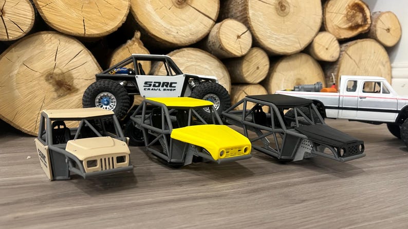 K&ouml;nnte beinhalten: Mehrere Miniatur-Gel&auml;ndewagen werden gezeigt, darunter ein wei&szlig;-schwarzer Rock Crawler mit dem Text "SDRC Crawl Shop", ein gelbbedachtes Buggy und ein wei&szlig;er Pick-up. Die Fahrzeuge stehen vor einem Hintergrund aus gestapelten Holzscheiten.