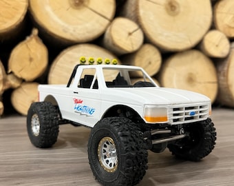 May include: A white toy pickup truck with large black tires and silver rims. The truck has the words "Miller Lightning" on the side and a row of yellow smiley faces on the roof. The truck is set against a backdrop of wooden logs.