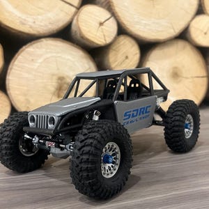 K&ouml;nnte beinhalten: Ein silbernes und schwarzes SDRC Crawl Shop Modell-Gel&auml;ndefahrzeug mit gro&szlig;en, schwarzen Reifen. Das Modell hat einen &Uuml;berrollk&auml;fig und der Text "SDRC Crawl Shop" ist auf der Seite sichtbar. Der Hintergrund zeigt gestapelte Holzscheite.