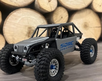 May include: A silver and black SDRC Crawl Shop model off-road vehicle with large, black tires. The model has a roll cage and the text "SDRC Crawl Shop" is visible on the side. The background features stacked wooden logs.