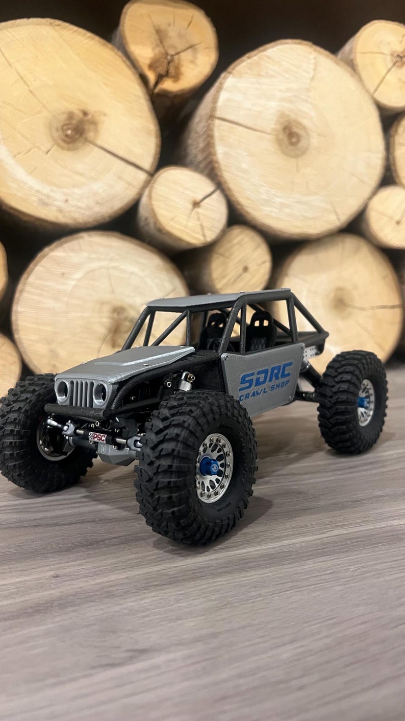 K&ouml;nnte beinhalten: Ein silbernes und schwarzes SDRC Crawl Shop Modell-Gel&auml;ndefahrzeug mit gro&szlig;en, schwarzen Reifen. Das Modell hat einen &Uuml;berrollk&auml;fig und der Text "SDRC Crawl Shop" ist auf der Seite sichtbar. Der Hintergrund zeigt gestapelte Holzscheite.