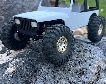 May include: White and black toy jeep with gold wheels. The jeep is sitting on a gray, textured rock.