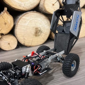 K&ouml;nnte beinhalten: Ein schwarzer und silberner ferngesteuerter Crawler mit gro&szlig;en, grobstolligen Reifen. Der Crawler hat ein detailliertes Fahrgestell und eine Karosserie, mit dem Text "SARC CRAWL SHOP" sichtbar. Der Hintergrund zeigt einen Stapel Holzscheite.