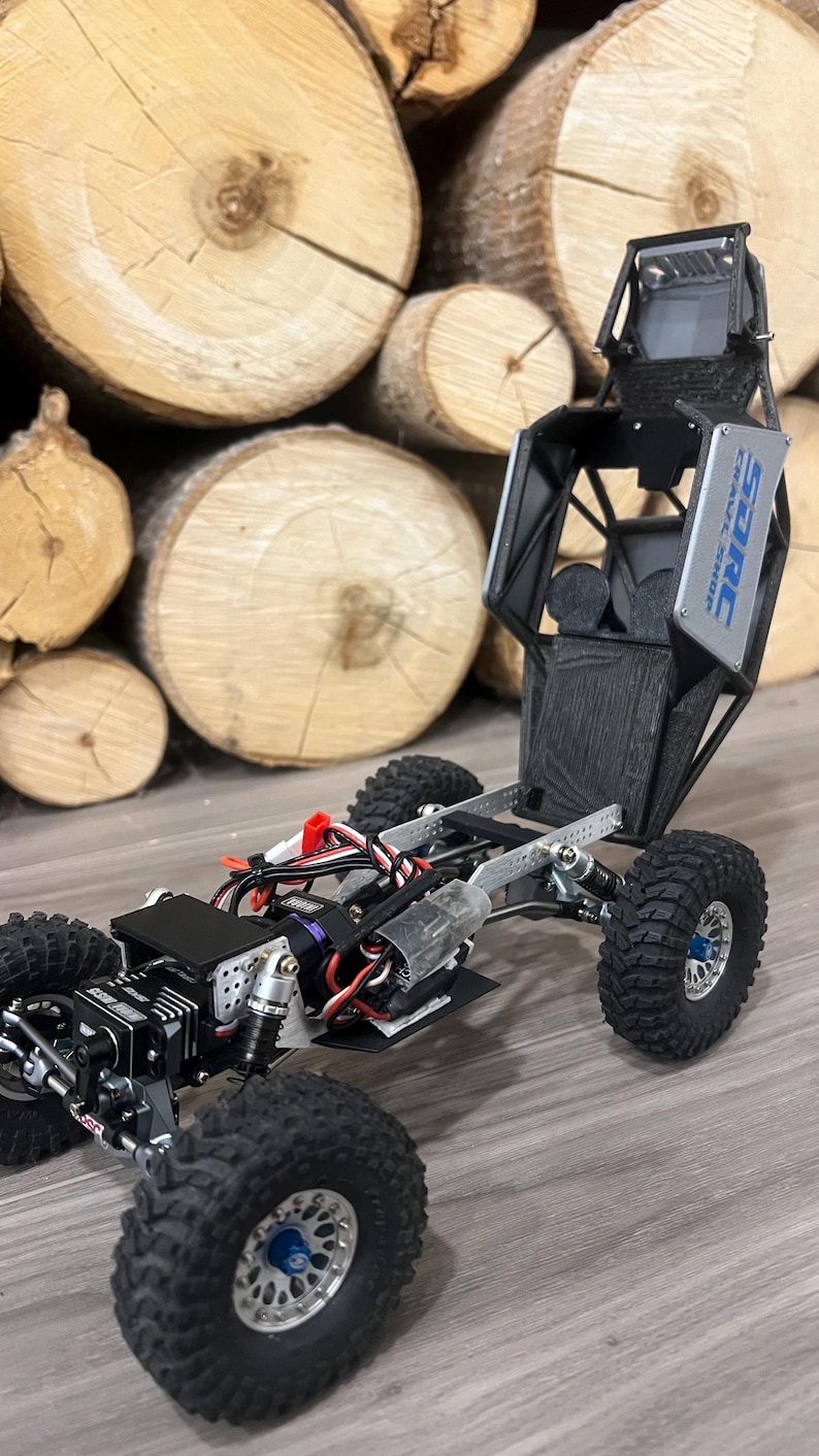 K&ouml;nnte beinhalten: Ein schwarzer und silberner ferngesteuerter Crawler mit gro&szlig;en, grobstolligen Reifen. Der Crawler hat ein detailliertes Fahrgestell und eine Karosserie, mit dem Text "SARC CRAWL SHOP" sichtbar. Der Hintergrund zeigt einen Stapel Holzscheite.