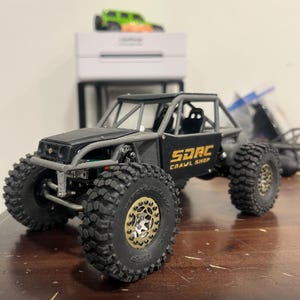 K&ouml;nnte beinhalten: Ein schwarzes und graues SDRC Crawl Shop Modellauto mit gro&szlig;en, grobstolligen Reifen. Der Truck hat einen &Uuml;berrollk&auml;fig und goldfarbene Raddetails. Ein gr&uuml;nes und orangefarbenes Spielzeugauto steht auf einer wei&szlig;en Box im Hintergrund.