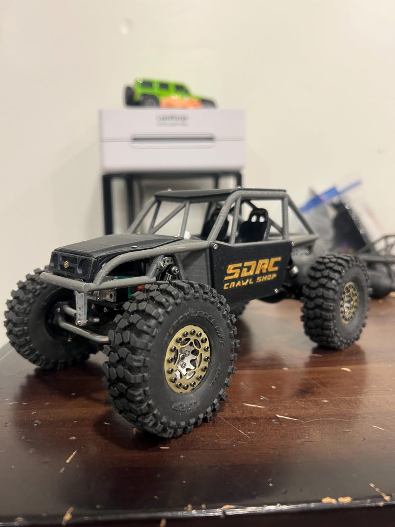 K&ouml;nnte beinhalten: Ein schwarzes und graues SDRC Crawl Shop Modellauto mit gro&szlig;en, grobstolligen Reifen. Der Truck hat einen &Uuml;berrollk&auml;fig und goldfarbene Raddetails. Ein gr&uuml;nes und orangefarbenes Spielzeugauto steht auf einer wei&szlig;en Box im Hintergrund.