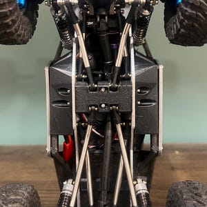 K&ouml;nnte beinhalten: Eine detaillierte Unteransicht eines kleinen, schwarz-silbernen ferngesteuerten Rock Crawlers. Das Modell verf&uuml;gt &uuml;ber gro&szlig;e, grobstollige Reifen, Federungskomponenten und ein komplexes Chassis-Design. Die R&auml;der sind schwarz mit blauen Akzenten.