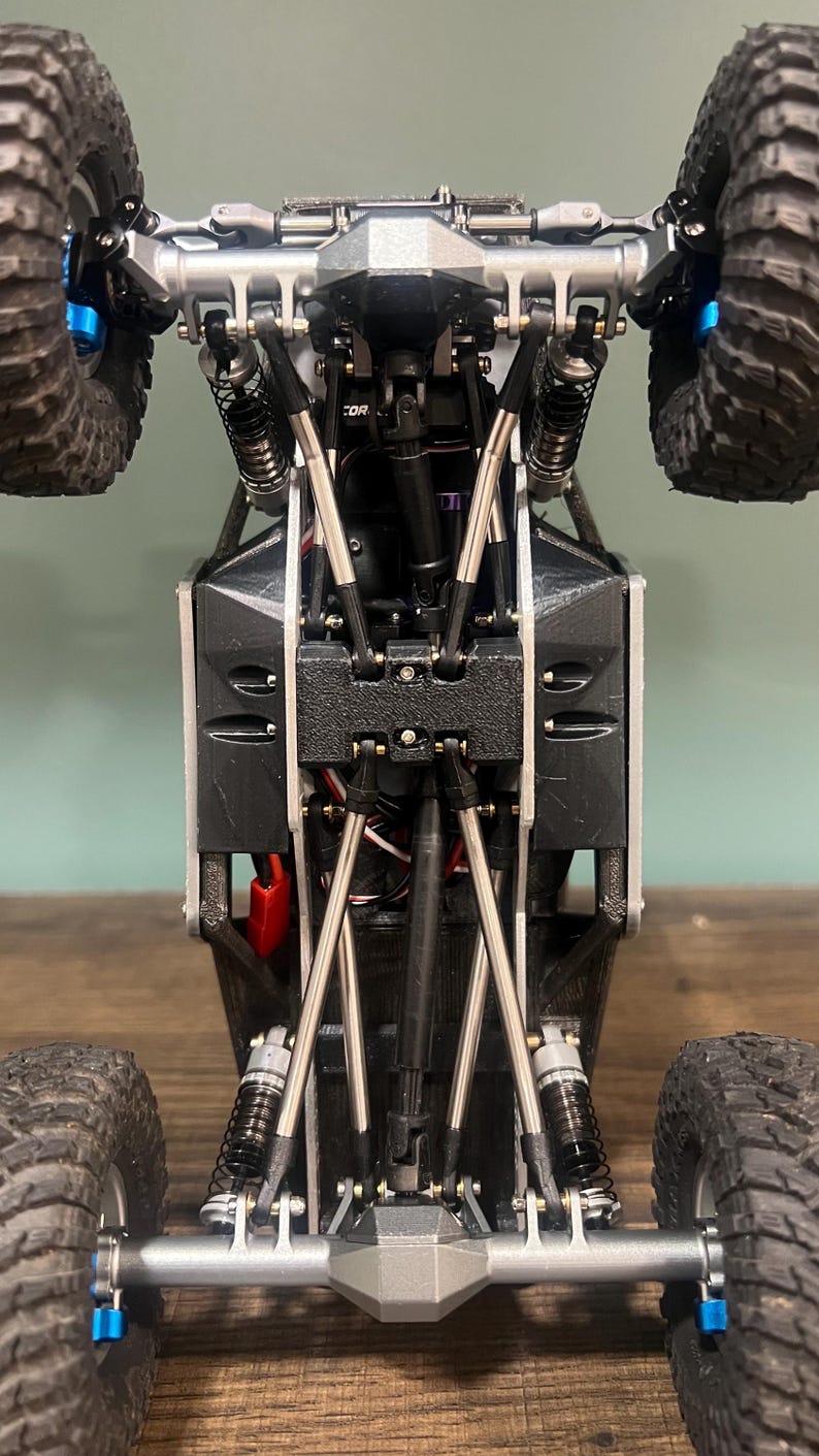 K&ouml;nnte beinhalten: Eine detaillierte Unteransicht eines kleinen, schwarz-silbernen ferngesteuerten Rock Crawlers. Das Modell verf&uuml;gt &uuml;ber gro&szlig;e, grobstollige Reifen, Federungskomponenten und ein komplexes Chassis-Design. Die R&auml;der sind schwarz mit blauen Akzenten.