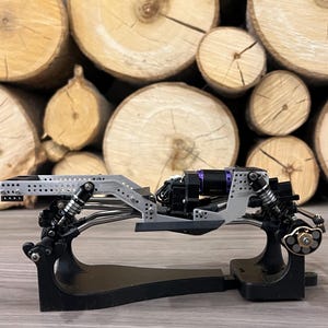 May include: A detailed view of a model car chassis, featuring a silver frame with black and purple components. The chassis is mounted on a black stand, showcasing the intricate design. The background is a stack of wooden logs.