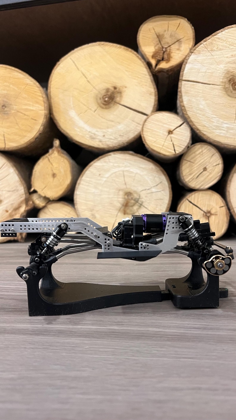 May include: A detailed view of a model car chassis, featuring a silver frame with black and purple components. The chassis is mounted on a black stand, showcasing the intricate design. The background is a stack of wooden logs.